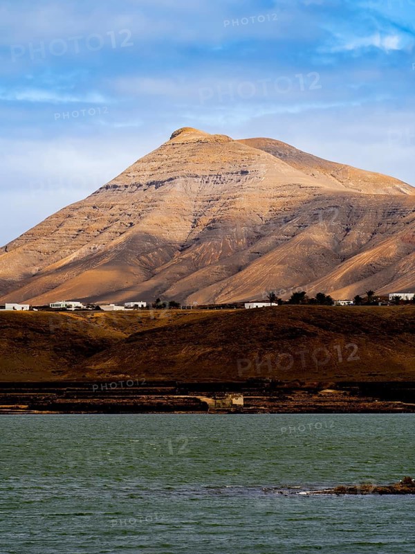 Majestic volcanic peak in Timanfaya National Park, Lanzarote, set against a vibrant blue sky with a serene foreground of water, showcasing dramatic geological formations