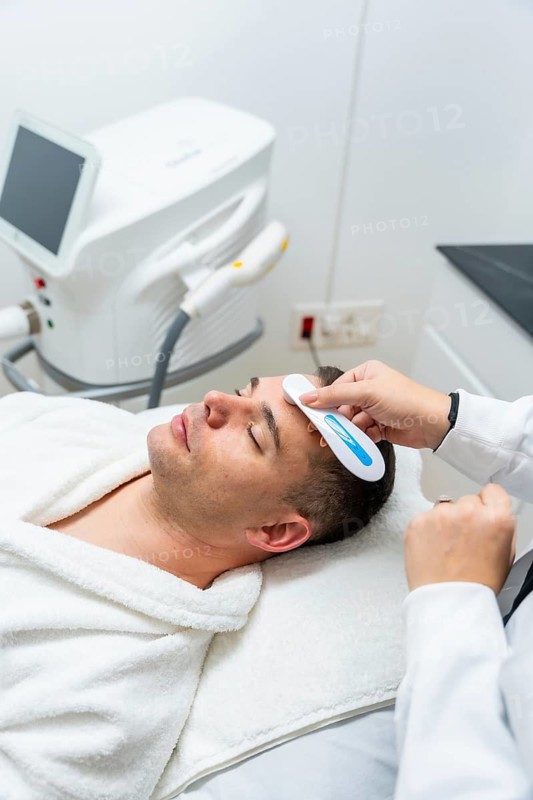 Male client relaxing wrapped in a white robe on a treatment bed while a professional cosmetologist performs a modern facial skincare procedure using a specialized device at a modern aesthetic clinic