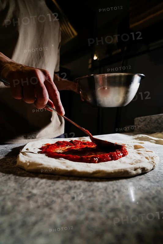 A person spreading fresh tomato sauce on a pizza dough in a dimly lit kitchen. The focus is on the process of creating a delicious homemade pizza from scratch