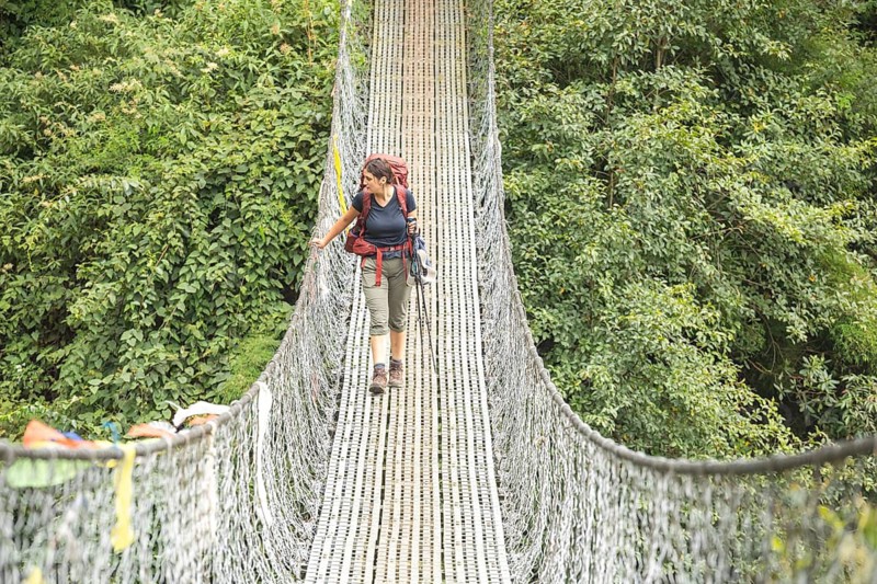 A woman treks across a suspension bridge surrounded by lush green foliage in the Everest region. Equipped with trekking gear, she embarks on a thrilling adventure