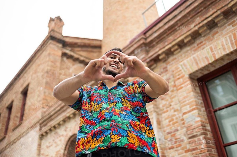 A cheerful man wearing a vibrant tropical shirt forms a heart shape with his hands while smiling. Captured from a low angle against a historic brick building, the image radiates positivity and joy