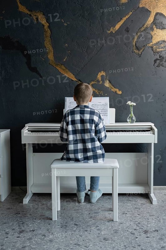 A young boy in casual attire sits on a bench, focused intently on playing a white electric piano in a stylish room. Sheet music and a small flower vase decorate the piano