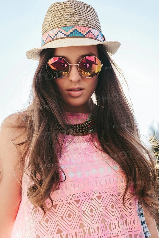 A young woman embodies summer style in a straw hat and stylish sunglasses. Her pink patterned top and statement necklace highlight a chic fashion look under the sunny sky
