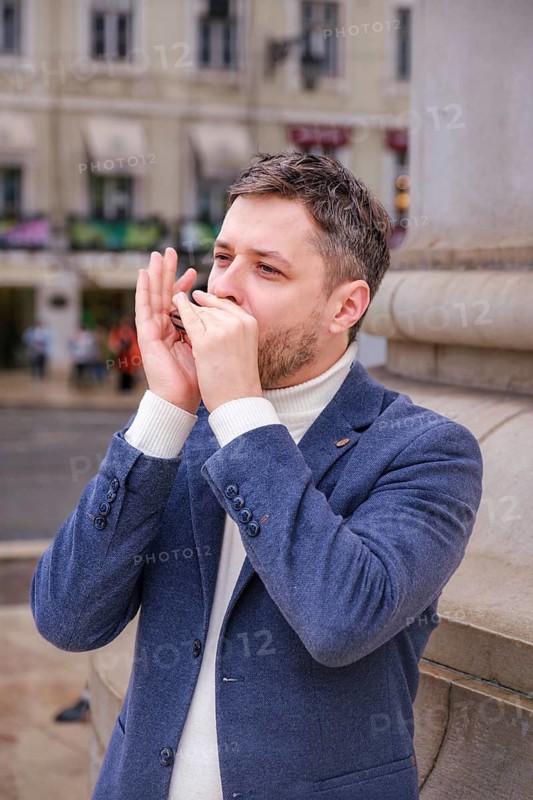 Man musician in a blue blazer and white sweater playing blues on a harmonica with eyes closed, standing outdoors in city street