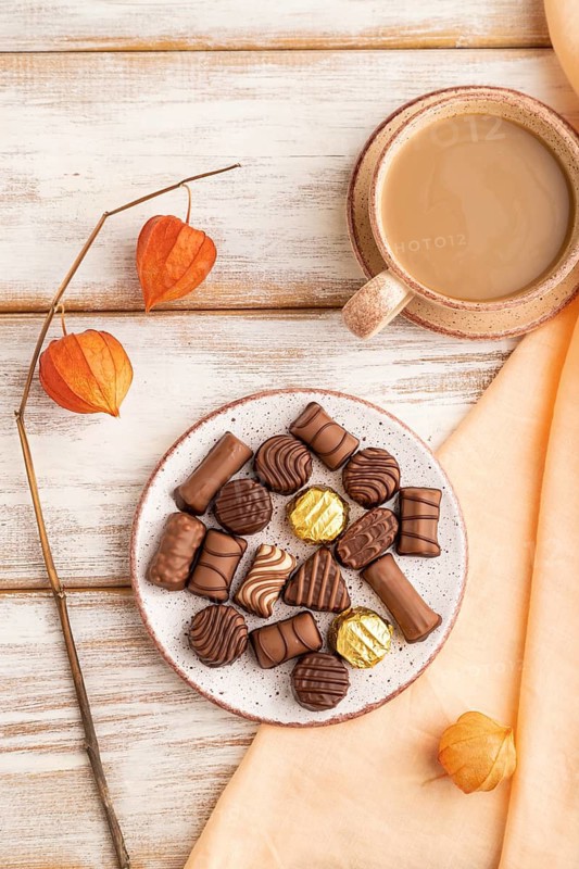 Chocolate candies with cup of coffee and physalis flowers on a white wooden background and orange textile. top view, flat lay, close up