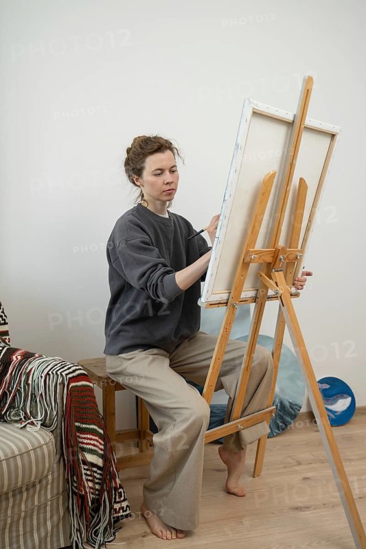 A focused artist paints on a canvas at an easel in a cozy home studio. The room exudes creativity with a striped blanket and art supplies, showcasing a minimalist aesthetic