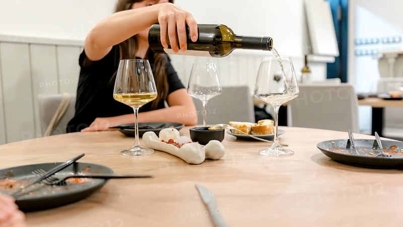 A woman pours white wine into a glass at a restaurant dinner setting. The table is elegantly set with empty plates and glasses, creating a cozy dining atmosphere