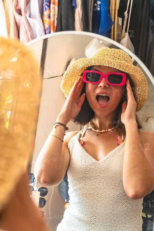 Young woman surprised by her reflection wearing pink sunglasses and straw hat in a clothing store mirror in siargao island, philippines