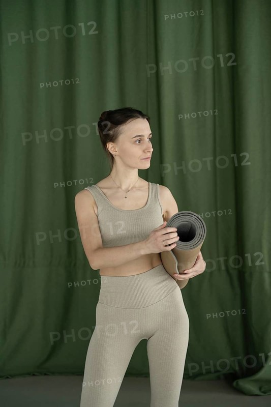 Woman in beige fitness attire holding a rolled yoga mat, set against a green curtain backdrop. Ideal for themes of wellness, fitness, and relaxation