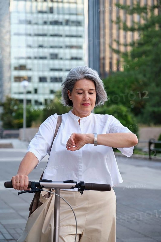 Stylish mature woman in urban setting glancing at her watch while holding a scooter. She is wearing a white blouse and appears focused and content outdoors