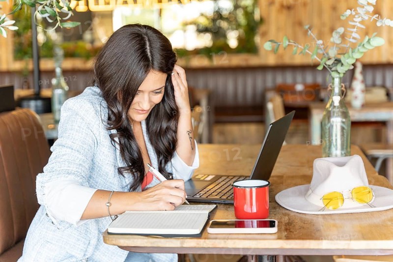 A woman writes in a notebook while using a laptop at a cozy cafe. Surrounded by a cup, hat, and sunglasses on a wooden table, she embodies a blend of work and relaxation