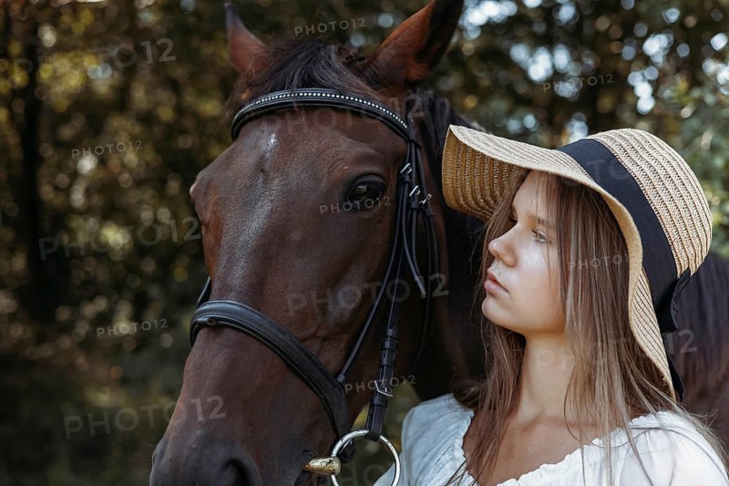 A young girl with long hair and a wide brimmed straw hat stands closely next to a brown horse surrounded by a peaceful forest backdrop, evoking a serene, rustic vibe