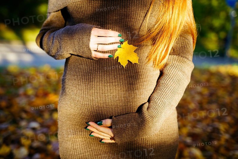 A serene image of a beautiful pregnant woman with long red hair holding a yellow autumn leaf. She stands amidst a warm, golden-hued fall backdrop, symbolizing growth and change