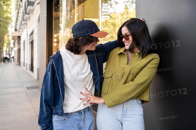 A happy lesbian couple embraces while walking down a sunny city street. They appear relaxed and joyful, sharing a warm moment together against a backdrop of urban architecture