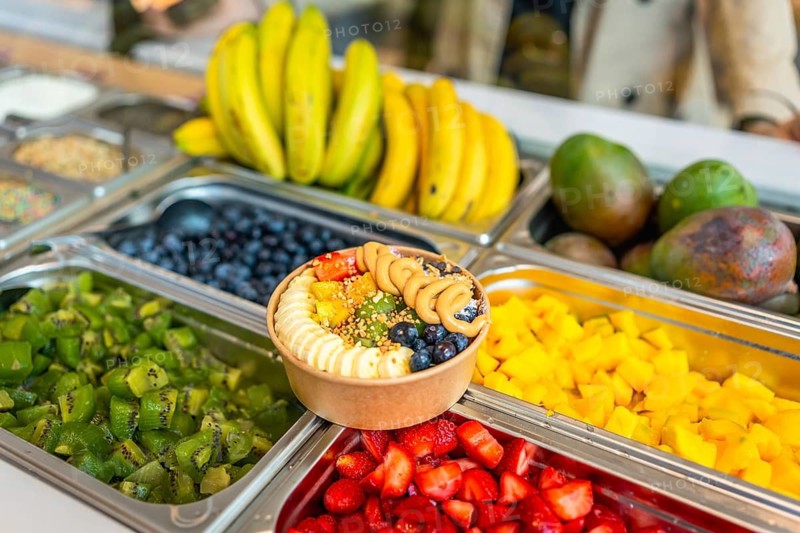 Acai smoothie bowl topped with bananas, blueberries, strawberries, kiwi, mango, nuts, and peanut butter, presented at a vibrant self service fruit bar