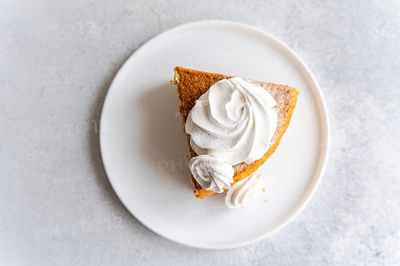 From above a slice of homemade honey cake topped with a swirl of vanilla zephyr sits elegantly on a white plate. Accompanied by a dollop of creamy sour cream sauce, this dessert is a visual delight