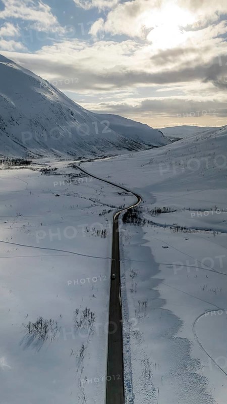Aerial view of a winding road cutting through the snow-covered Nordic landscape, surrounded by majestic mountains under a partly cloudy sky, capturing winter's serene beauty, Kvaløya, Troms, Norway