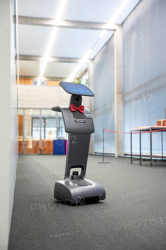 A robot-like device with a red ribbon stands on a carpeted floor in an illuminated office, interactive guided tours by robotic personalities from Robolutions, Pforzheim, Germany