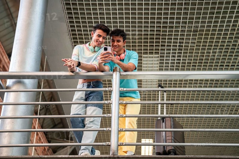 A joyful gay couple stands at a railing, sharing a moment and looking at a smartphone. They appear relaxed and happy, surrounded by luggage, suggesting travel or transit