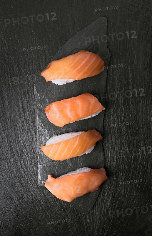 Four pieces of fresh salmon sushi arranged neatly on a textured black stone surface. The vibrant orange of the salmon contrasts with the dark background, emphasizing elegance