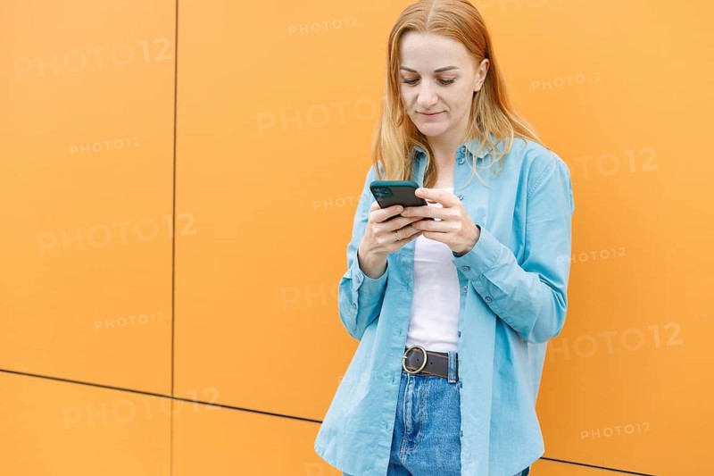 A woman in a casual blue shirt stands against a vibrant orange wall in Warsaw, focused on her smartphone The scene captures a modern urban lifestyle moment