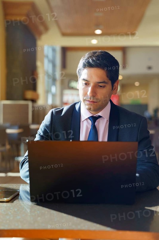 A young Chilean businessman in formal attire works diligently on his laptop in a modern cafe. Sunlight filters through large windows, creating a focused and professional ambiance