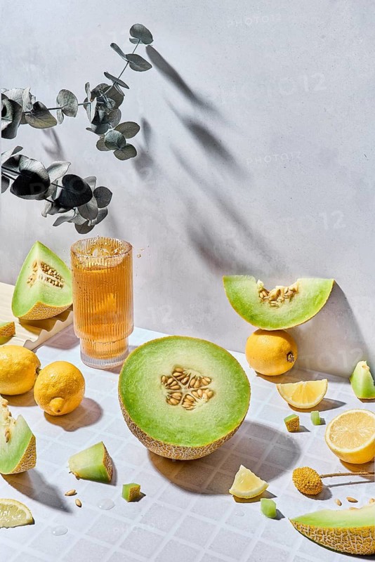 A vibrant display of fresh melons, lemons and fresh juice in a glass on a textured white background, highlighted by warm sunlight. The composition evokes fresh summer vibes and a light mood