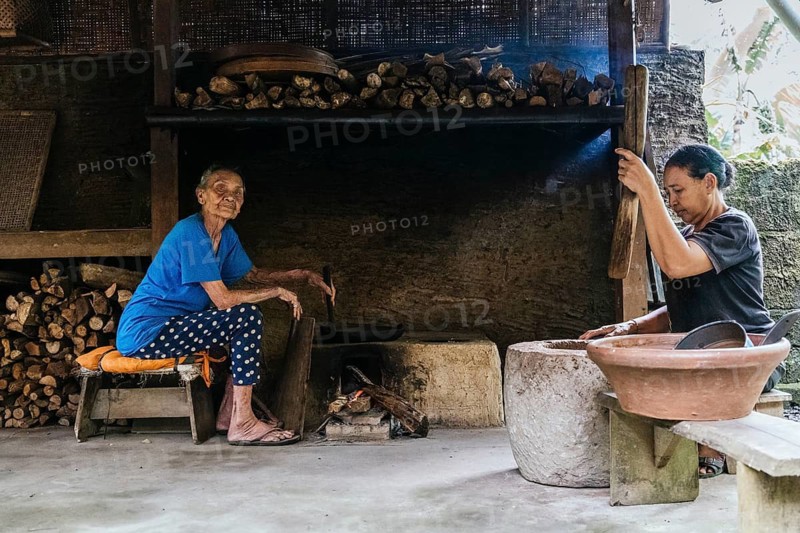 Two women work in a rustic kitchen, surrounded by firewood and traditional cookware. One prepares coffee while the other tends to the fire, exemplifying cultural culinary practices