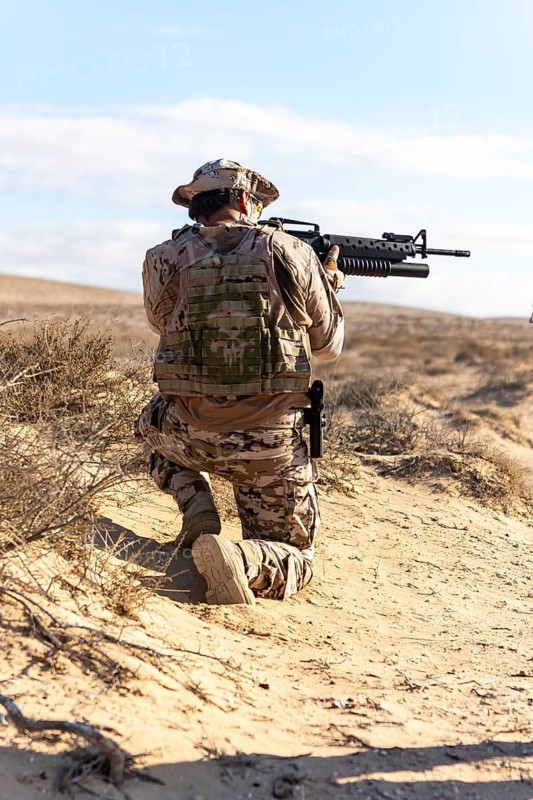 Soldier in full desert camouflage gear kneeling with firearm, ready for action. Training in open desert landscape under clear sky, representing military readiness and strategy