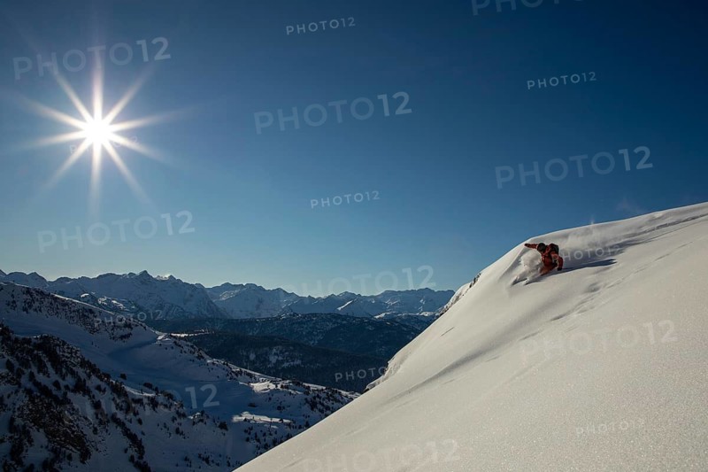 A skier glides down a steep, pristine snow-covered mountain slope under a clear blue sky The sun shines brightly, casting sharp shadows on the untouched snow