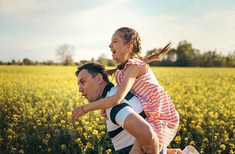 A joyful father carries his daughter on his back as they laugh together in a sunlit flower field. The warm golden light captures their happiness, connection, and playful family moment