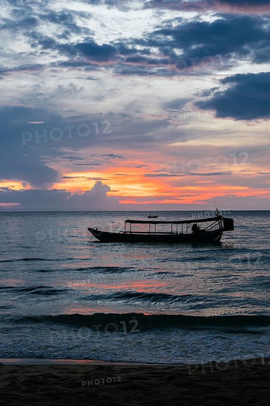 A traditional boat is silhouetted against a stunning sunset over the tranquil waters of Koh Rong Island, Cambodia. The sky is painted with hues of orange, pink, and purple