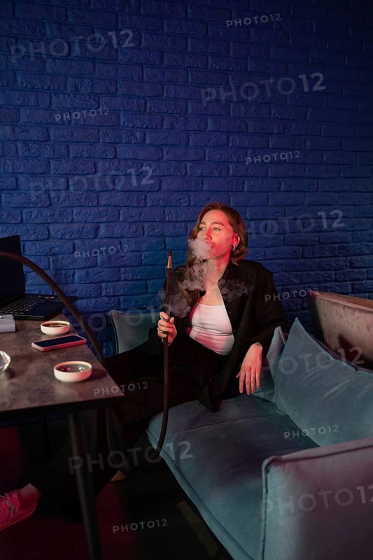 A woman enjoys a relaxed smoking session, sitting on a blue sofa with a hookah. The ambient red and blue lighting creates a calming atmosphere