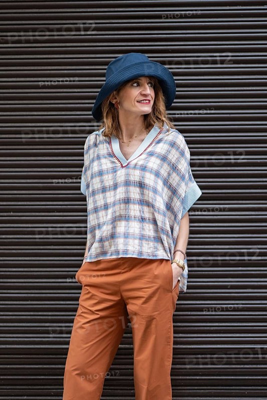 A stylish woman in a dark background, wearing a blue hat, plaid shirt, and orange pants, exuding relaxed elegance and modern fashion sense