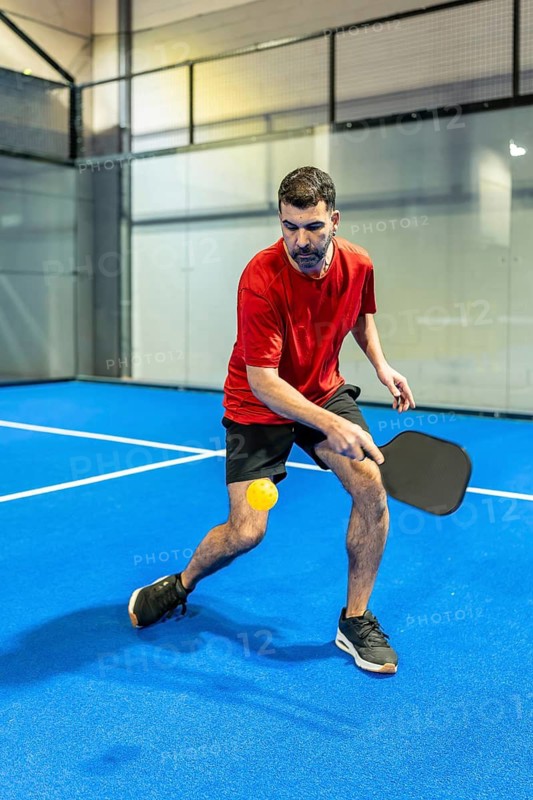 Male pickleball player intensely focusing on hitting the ball with a paddle during an indoor match on a blue court, showcasing action, agility and competitive spirit