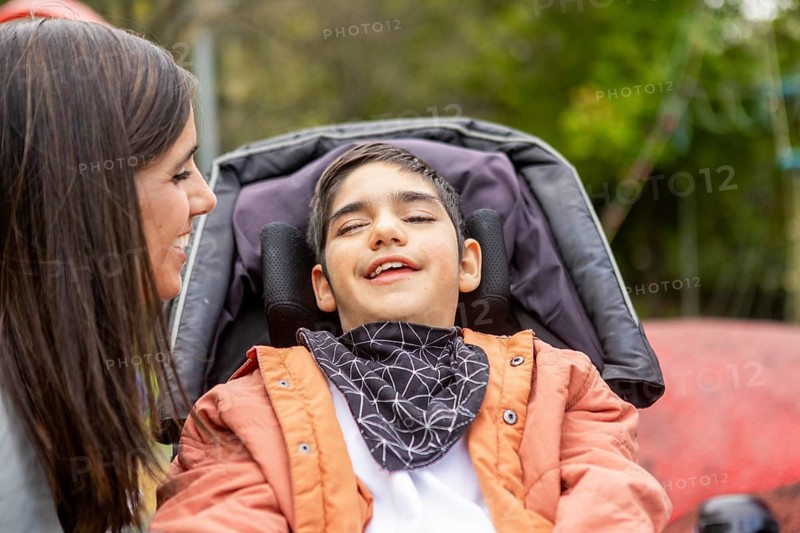A woman smiles warmly at a her child with cerebral palsy in a wheelchair, enjoying a sunny day in a playground. The loving interaction highlights care, support, and the joy of togetherness