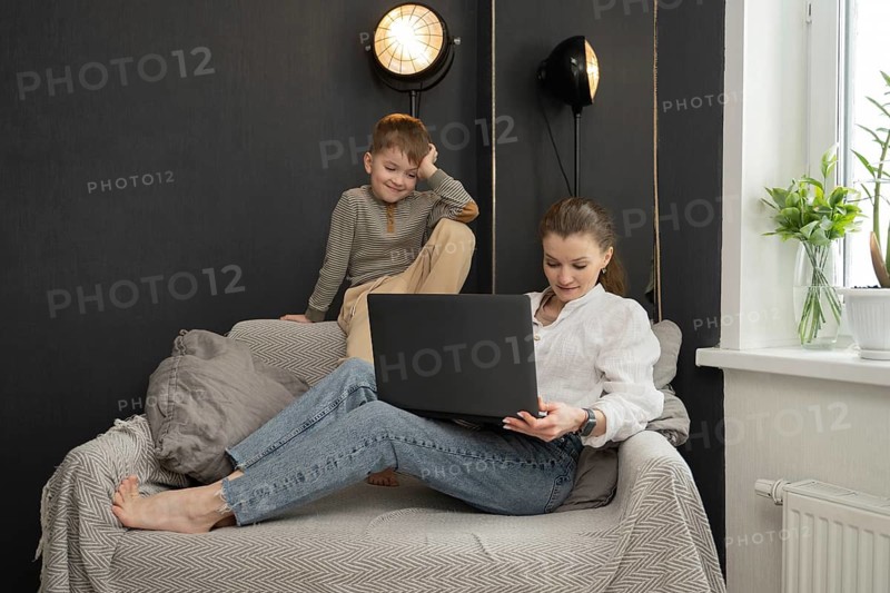 A mother lounges on a cozy sofa, working on her laptop, while her playful child sits nearby. The room features soft lighting and a decorative plant on the window sill