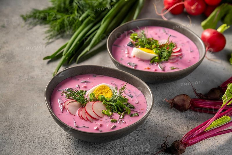 Two bowls of vibrant pink cold beetroot soup are garnished with sliced radishes, dill, green onions, and egg halves. Fresh beetroots and herbs surround the setup, enhancing the freshness