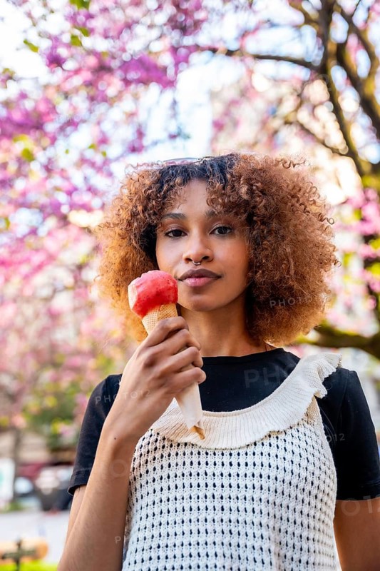 Stylish young woman savoring a strawberry ice cream cone against a backdrop of blooming pink trees in a springtime urban setting