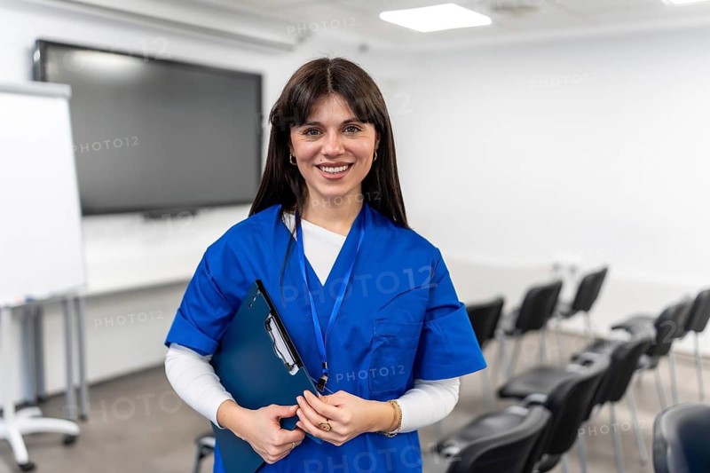 Young female medical professional wearing blue scrubs and id badge, smiling while holding a clipboard, standing confidently in an empty seminar room ready for a training session or class