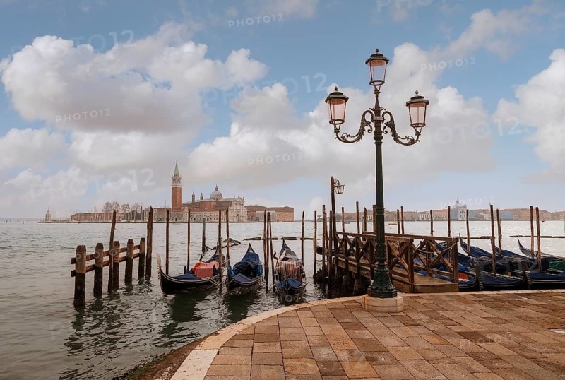 Picturesque Venetian scene with gondolas lined up along the dock and an ornate lamp post, set against a backdrop of historic architecture and a cloudy sky