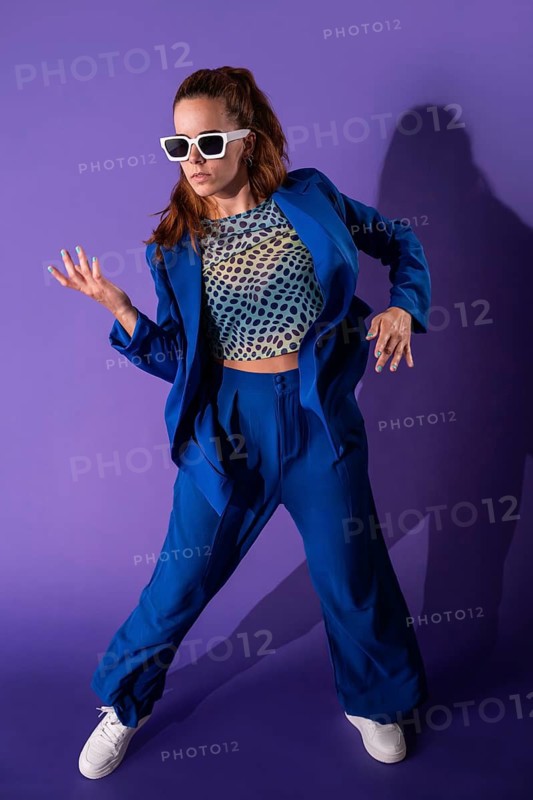 A young woman dancer striking a dynamic pose against a vibrant purple background. She displays energy and style, embodying the essence of modern dance in a colorful outfit