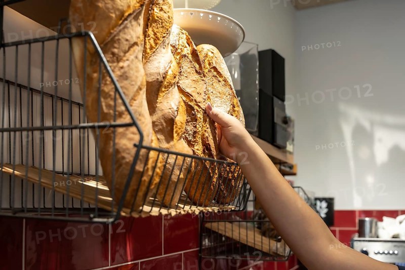 Customer hand selecting a freshly baked baguette from a wire basket, showcasing the artisanal bread production and authentic shopping experience in a local shop