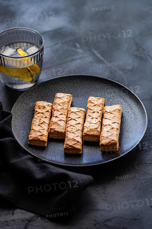 Crispy puff pastry sticks filled with sweet apricot jam, elegantly arranged on a sleek black plate. A refreshing glass of water with lemon slices complements this delightful treat