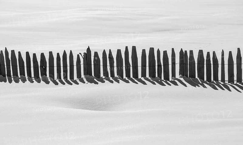 A monochrome image capturing a simple wooden fence casting long shadows across a pristine snow-covered landscape The minimalistic scene evokes tranquility and solitude