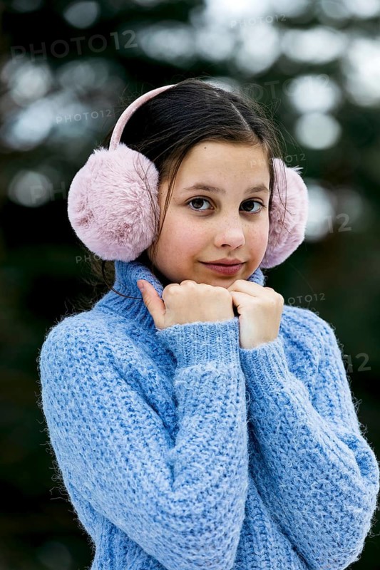 A young girl in a blue knitted sweater and pink earmuffs enjoys a snowy day. The cozy winter attire reflects warmth and comfort against a blurred snowy background