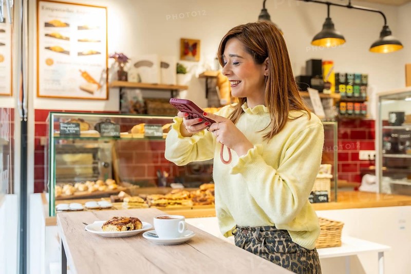 Woman capturing a delicious cinnamon roll and coffee with her smartphone, sharing a moment of leisure and showcasing the appealing breakfast pastry in a modern cafe setting
