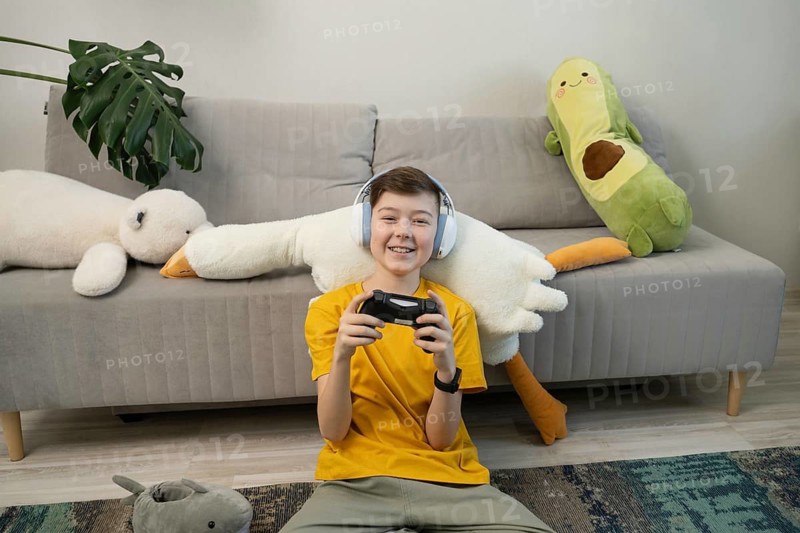 A cheerful boy wearing headphones plays video games with a controller in a cozy living room. Surrounded by plush toys, he sits on the floor, enjoying his leisure time
