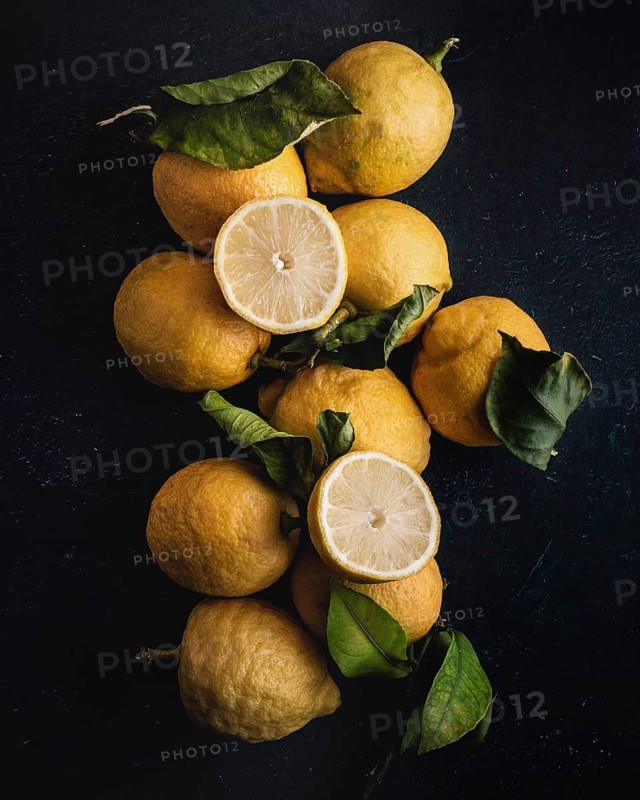 A rustic arrangement of fresh lemons, including a cut lemon revealing its juicy interior, sits atop a dark background. Bright green leaves add contrast to the yellow citrus