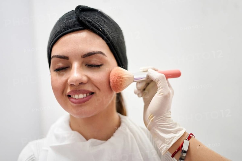 A beauty therapist applies makeup to a client's face in a modern salon. The setting emphasizes relaxation and professionalism with a clean environment